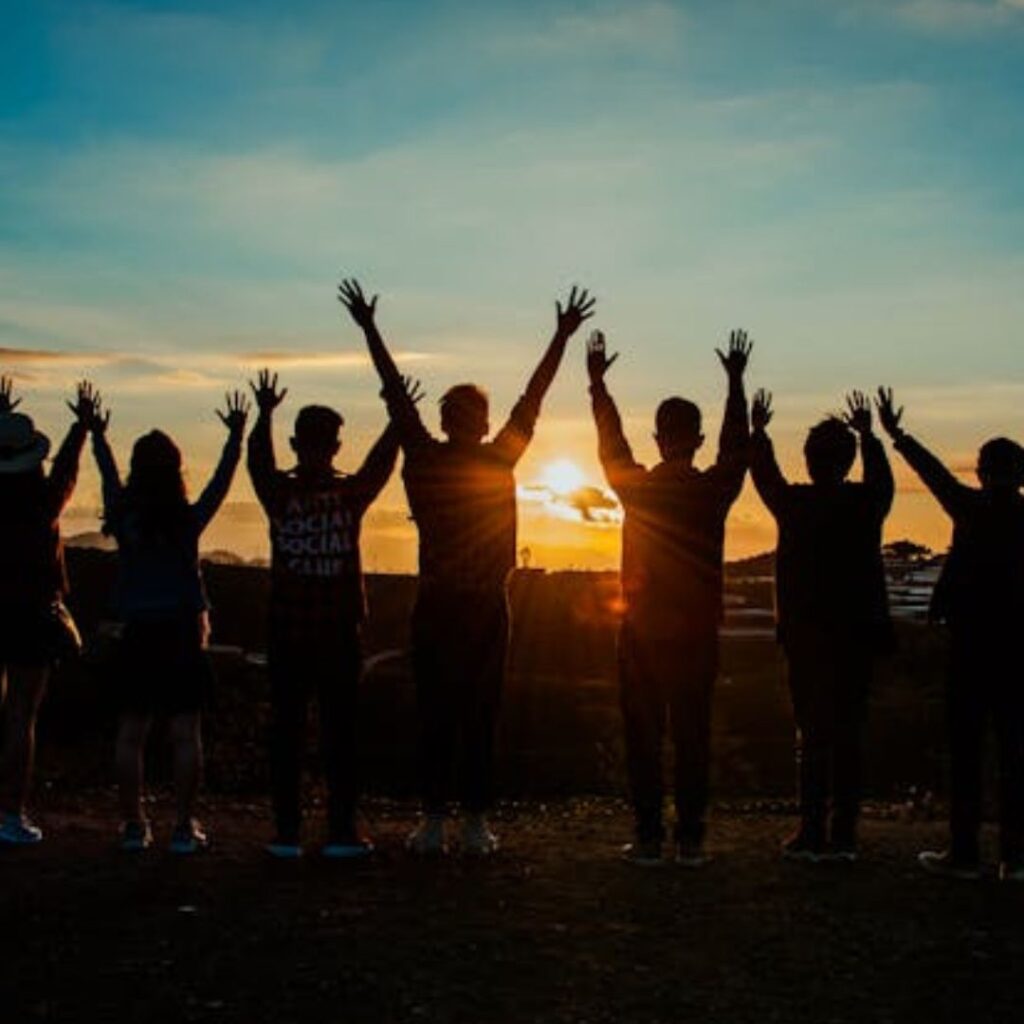 many individuals standing facing the sunset with hands raised