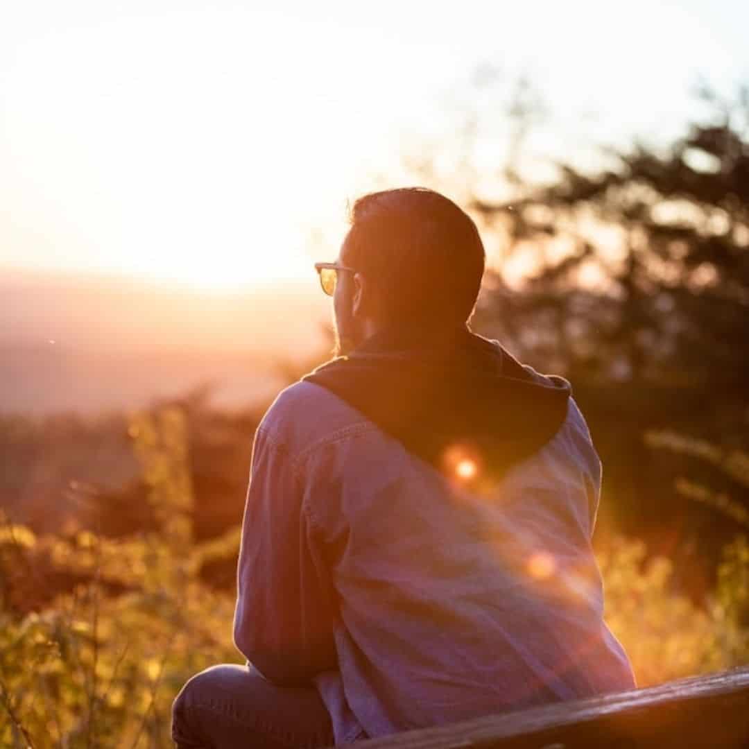 man sitting on bench out in nature, watching the sun rise in the distance
